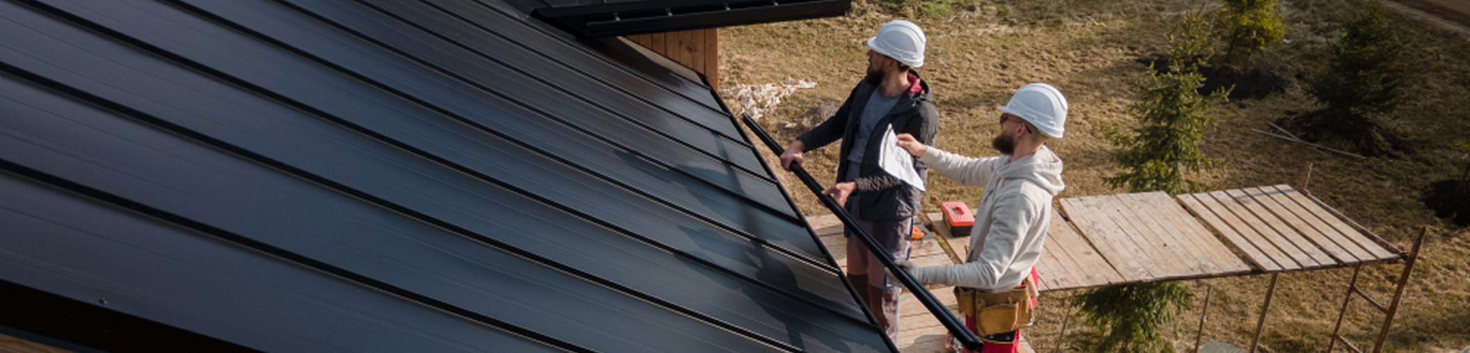 Two construction workers wearing white helmets inspecting a dark metal roof near wooden scaffolding.