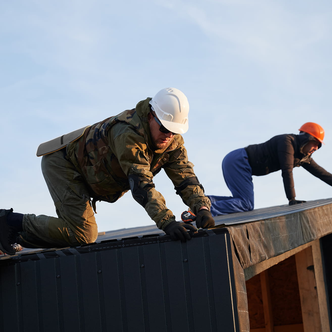 Two construction workers wearing helmets installing roofing panels on a sloped roof under a clear sky.