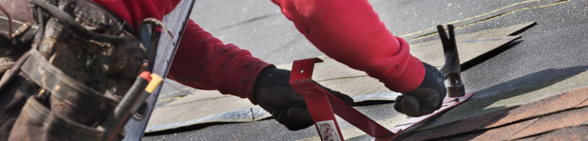 Roofer wearing black gloves and a red long sleeve shirt using a hammer to secure roofing shingles.