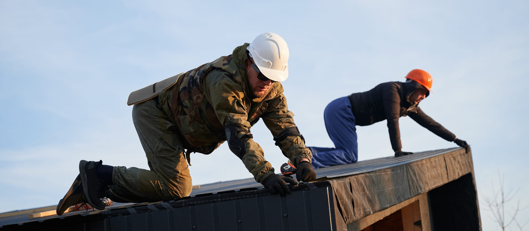 Two construction workers wearing safety helmets installing roofing materials on a building under clear sky.