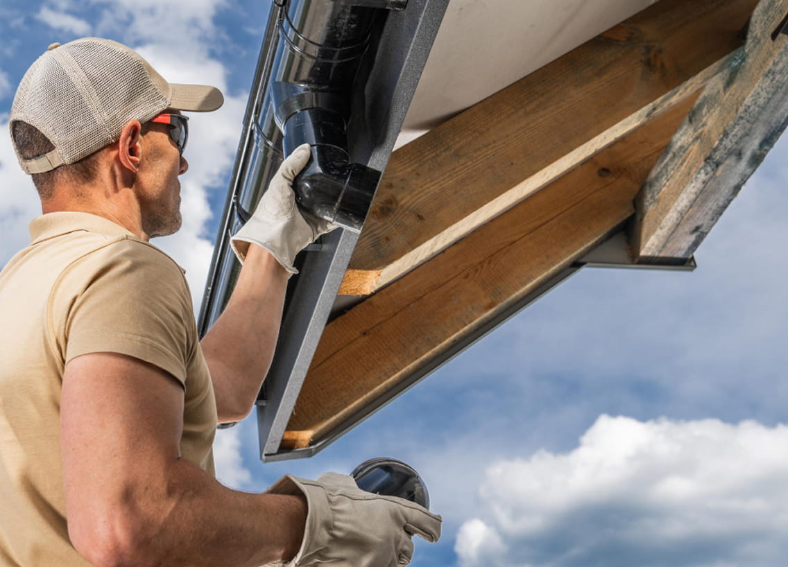 Man wearing gloves and cap installing or inspecting a black rain gutter on a wooden roof under a blue sky with clouds.