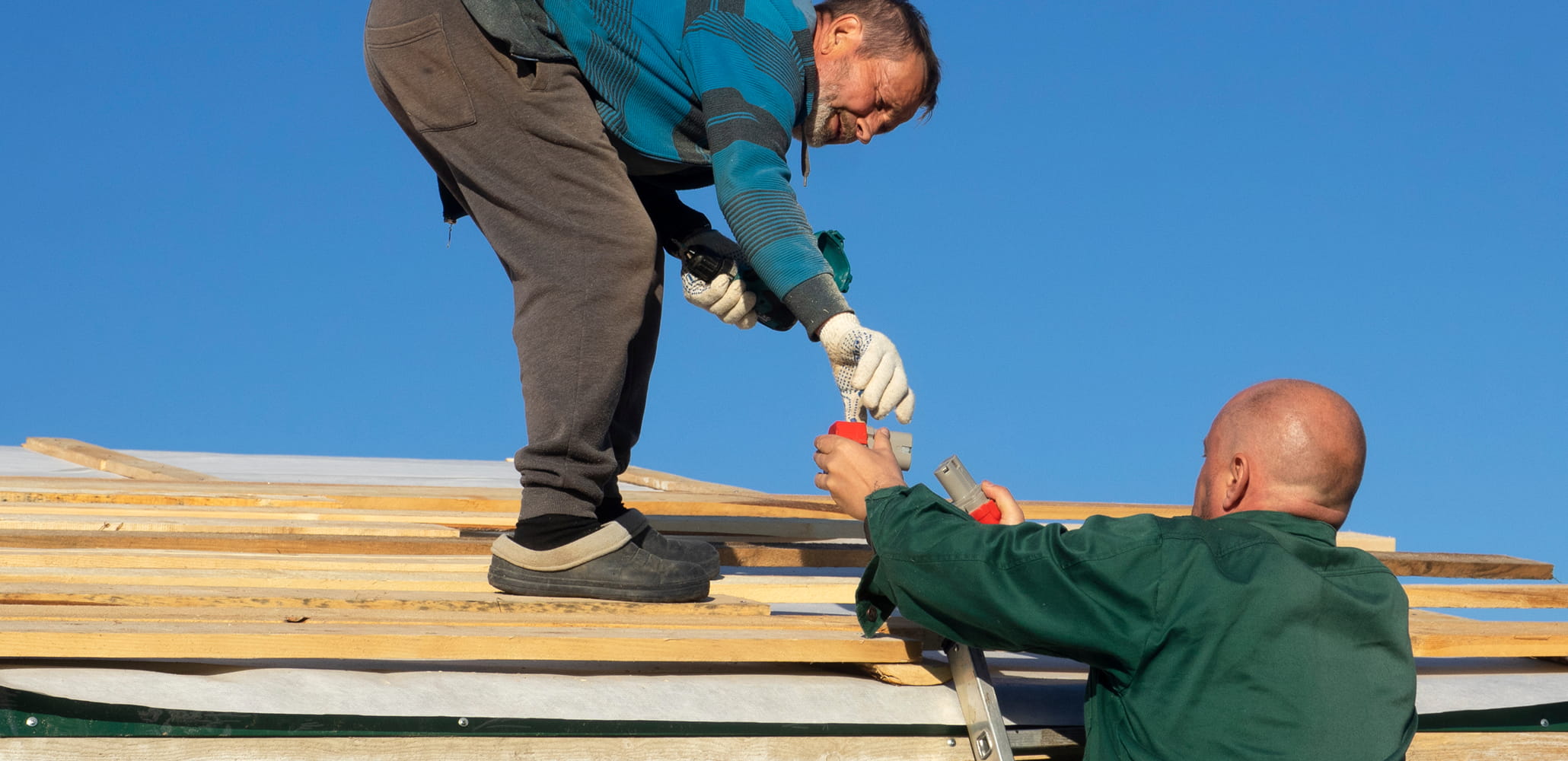 Two workers on a roof handing over a tool under a clear blue sky.