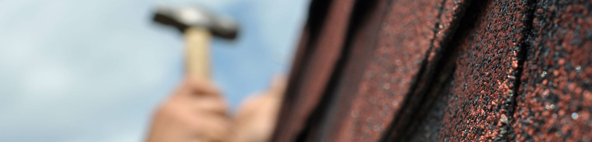 Close-up of a roof shingle on the right with a blurred hand holding a hammer in the background.