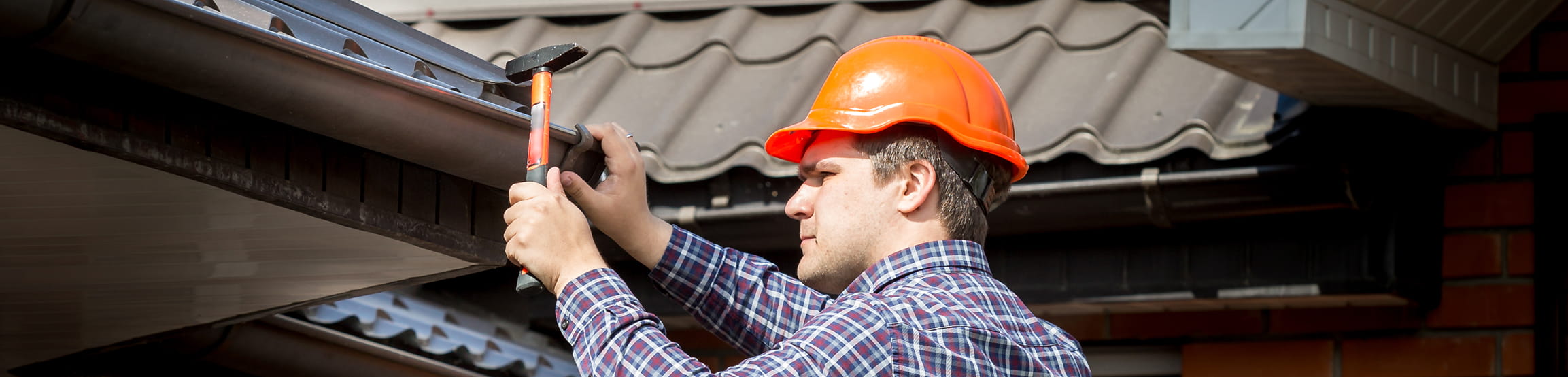Man wearing an orange hard hat and plaid shirt repairs a roof gutter using a hammer.