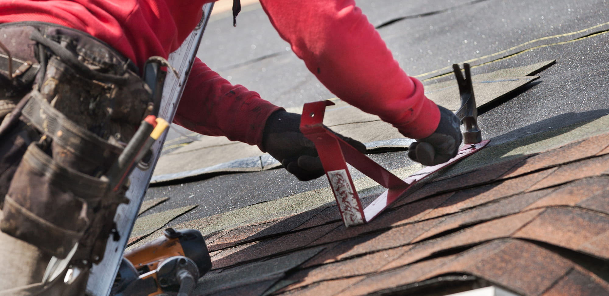 Roofer in red sweatshirt using a hammer and a red tool to install asphalt shingles on a roof.