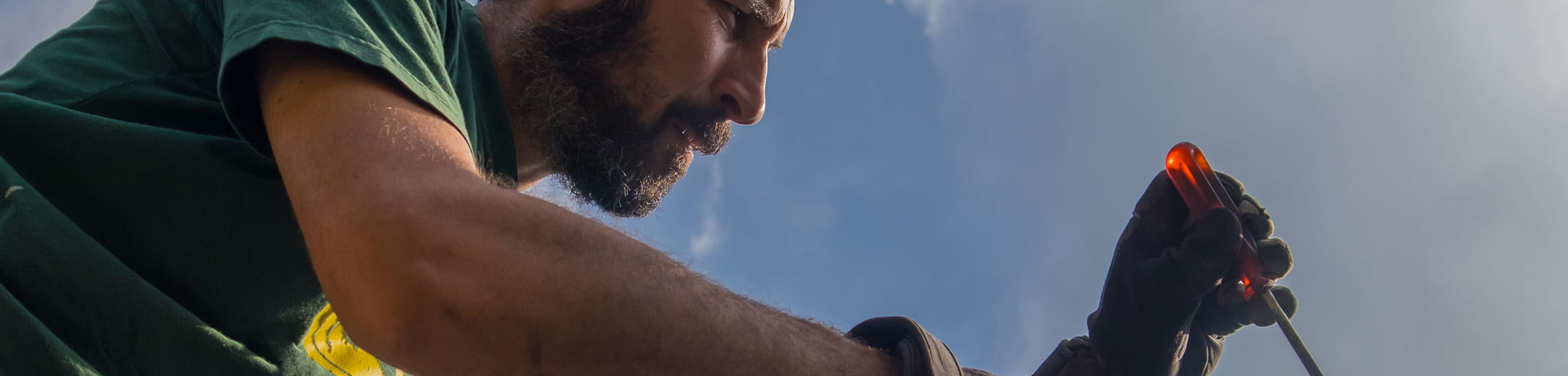 Man wearing gloves and holding a red screwdriver against a clear sky background.
