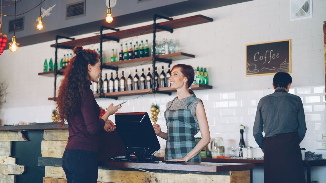Two women engaged at a coffee shop counter with a barista working in the background