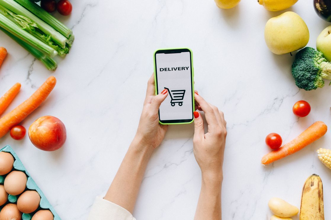 A person using a smartphone for grocery delivery surrounded by fresh fruits and vegetables