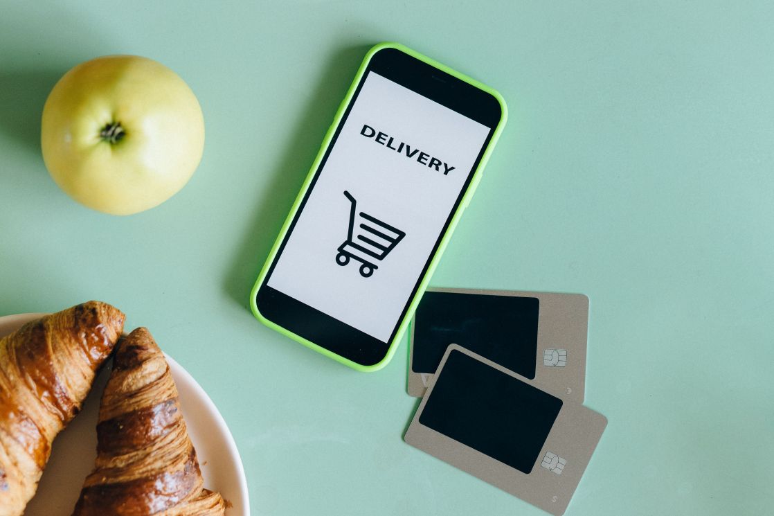 A smartphone with a delivery app next to an apple, croissants, and credit cards on a green surface