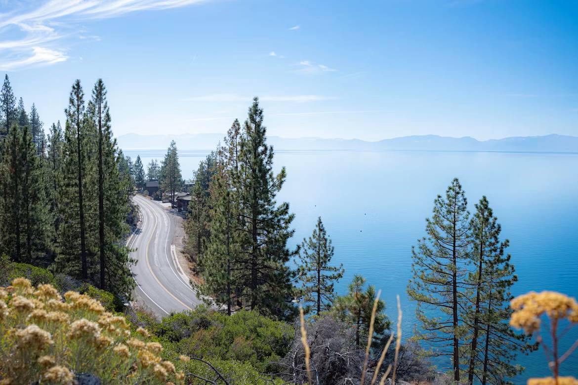 A picturesque empty road curving alongside a tranquil lake, framed by lush mountains under a clear sky.