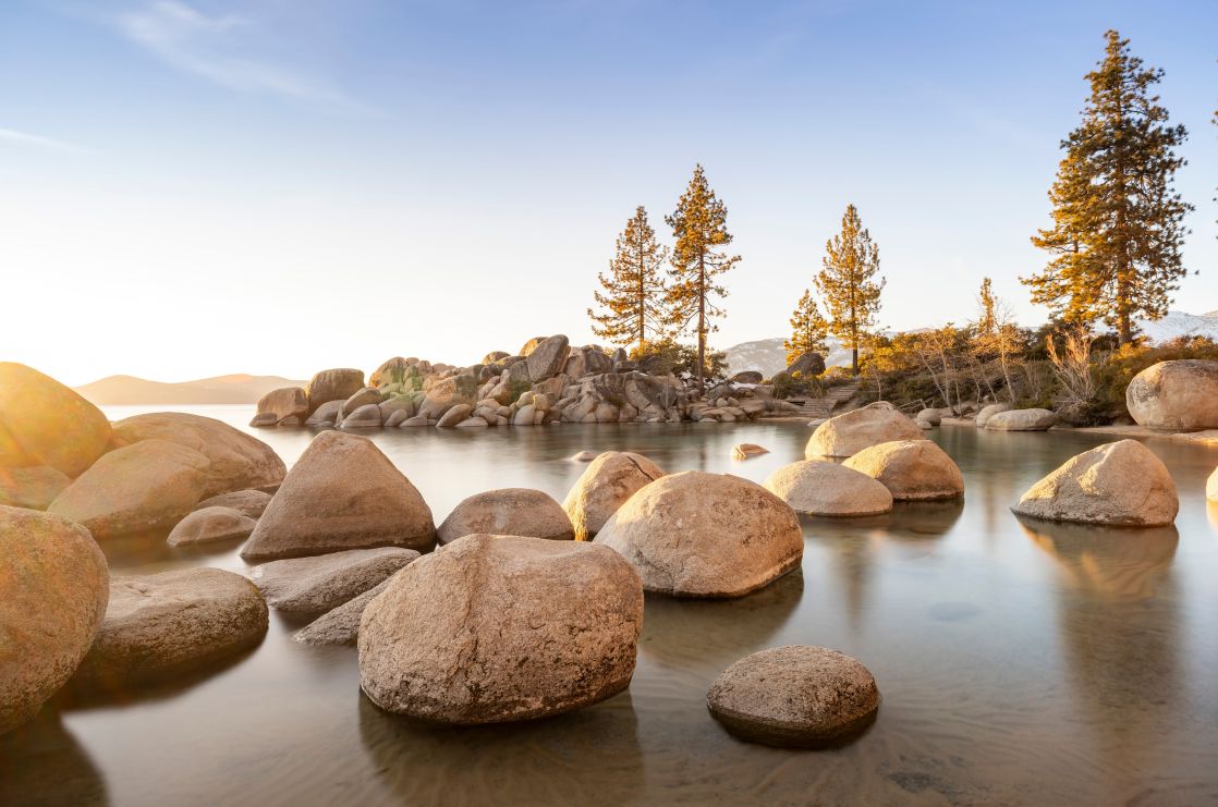 Peaceful scene of Lake Tahoe at sunset with rocks and trees reflecting in the calm waters.