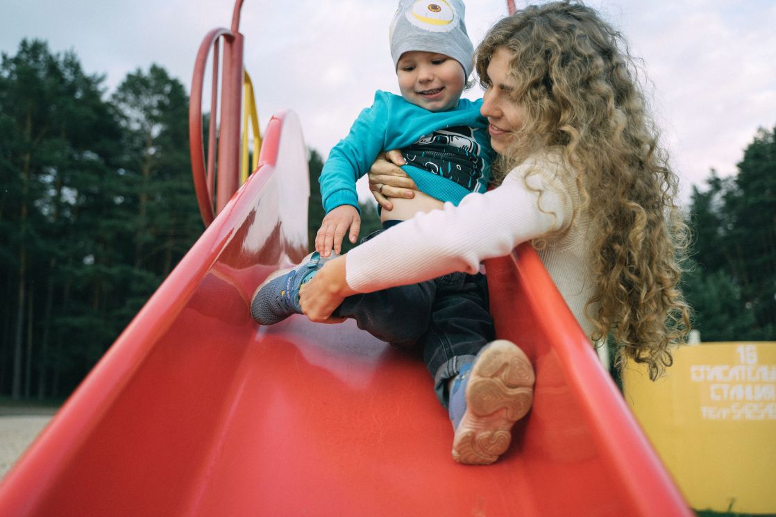 A happy mother helps her child slide down a red playground slide outdoors.