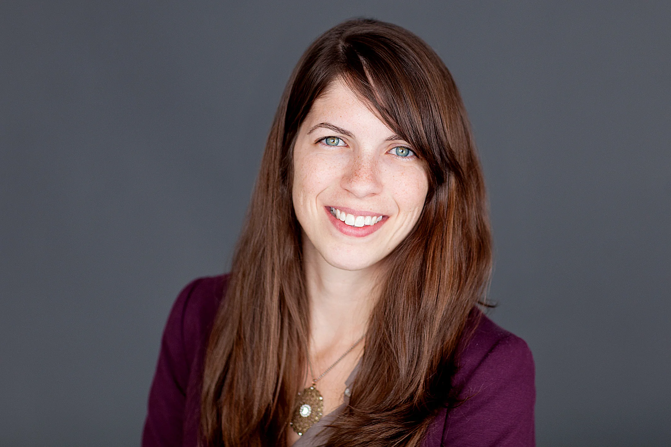 Smiling woman with long brown hair wearing a purple top and pendant necklace against gray background.