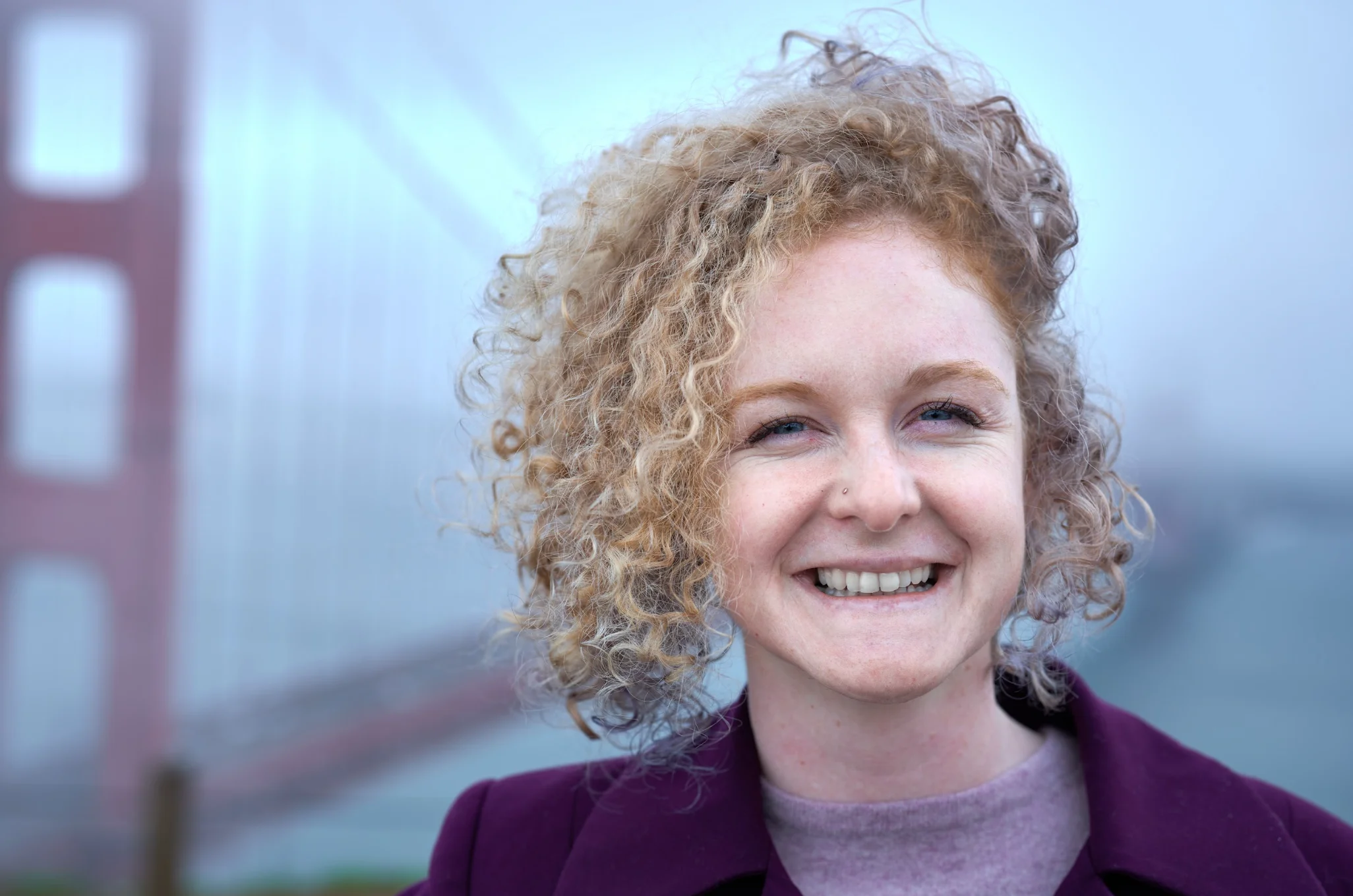 Smiling woman with curly blonde hair wearing purple jacket in front of a foggy Golden Gate Bridge.