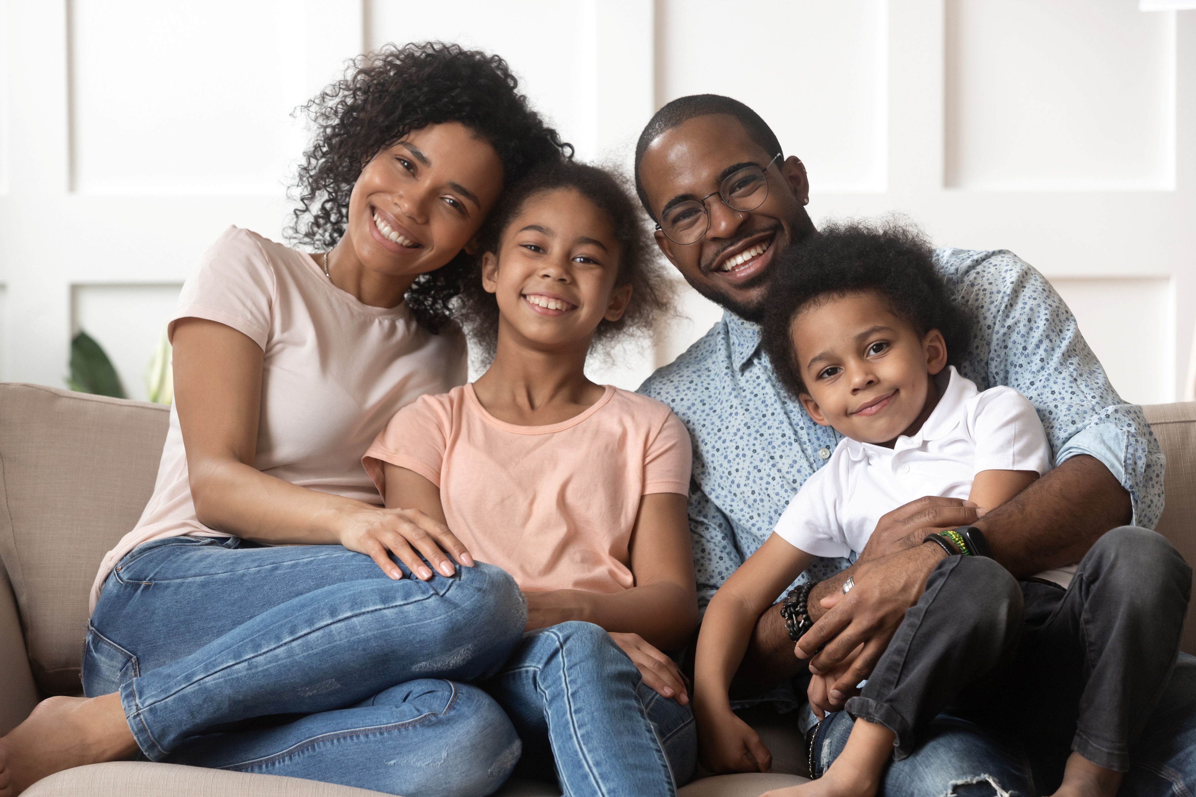 Happy family of four sitting on a couch smiling at the camera in a bright living room.