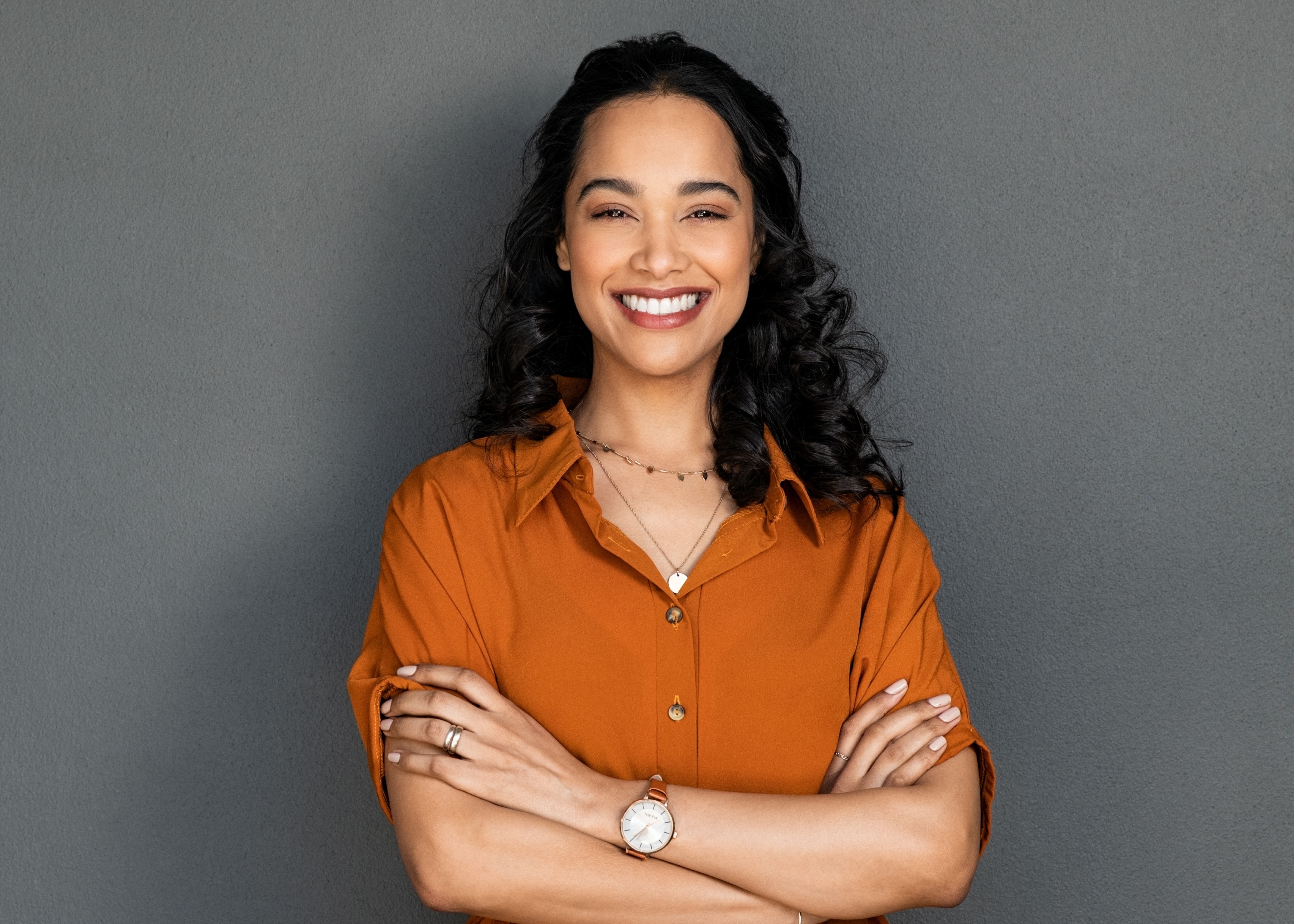 Smiling woman with curly hair in an orange shirt standing with arms crossed against a gray background.