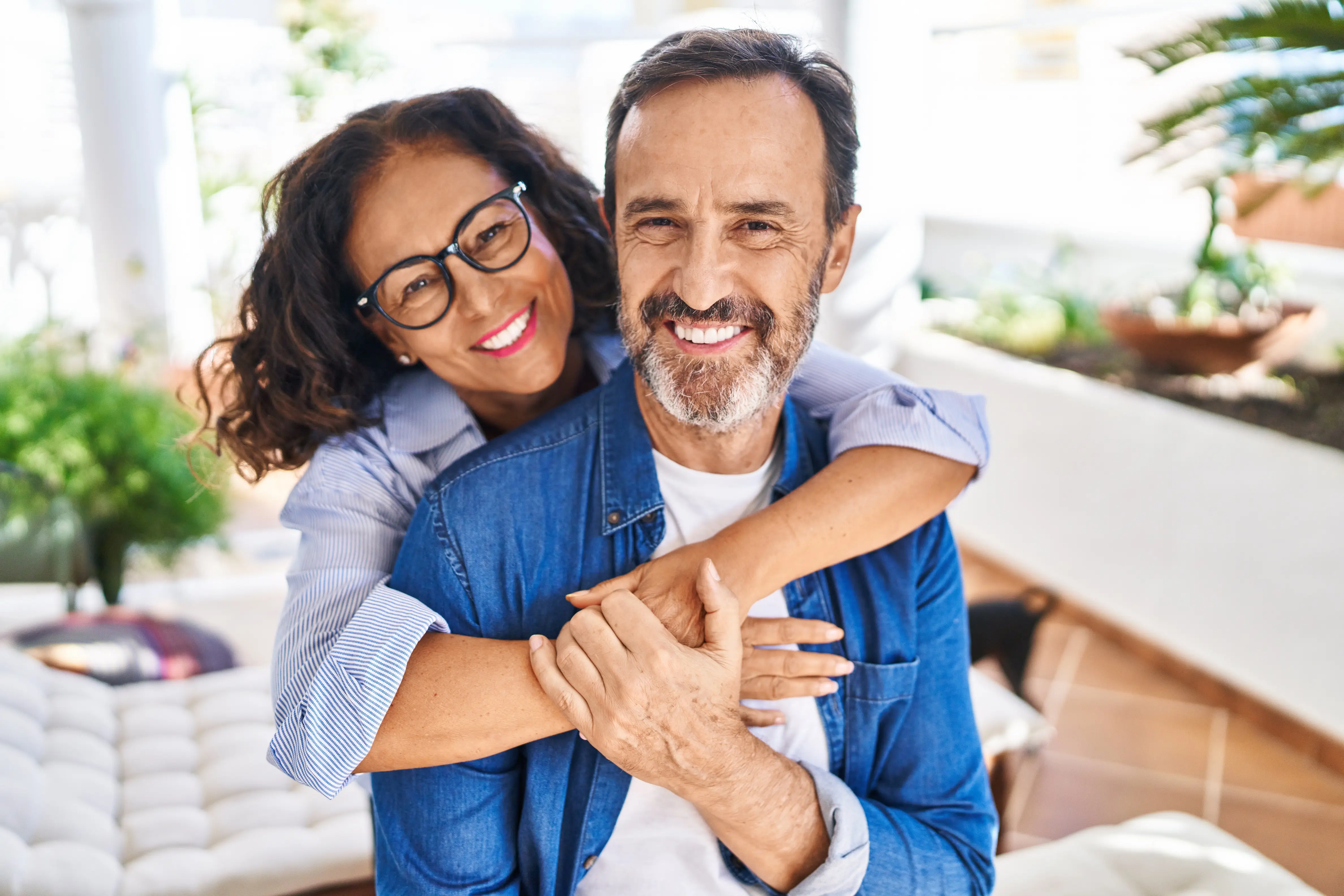 Smiling middle-aged couple hugging lovingly indoors with plants in the background.