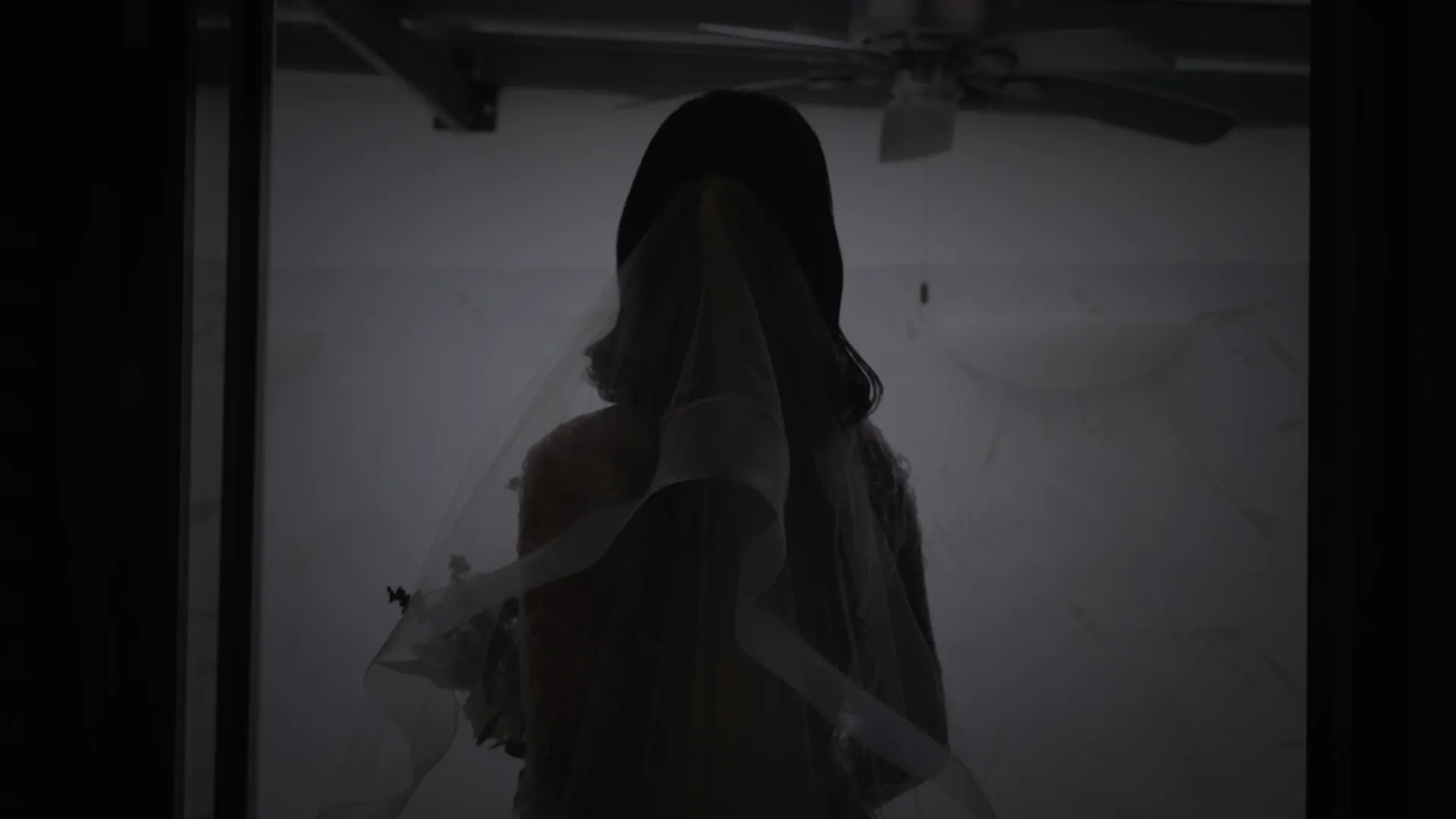 Silhouette of a bride with a veil holding a bouquet, standing indoors in front of a light-colored wall with a ceiling fan above.