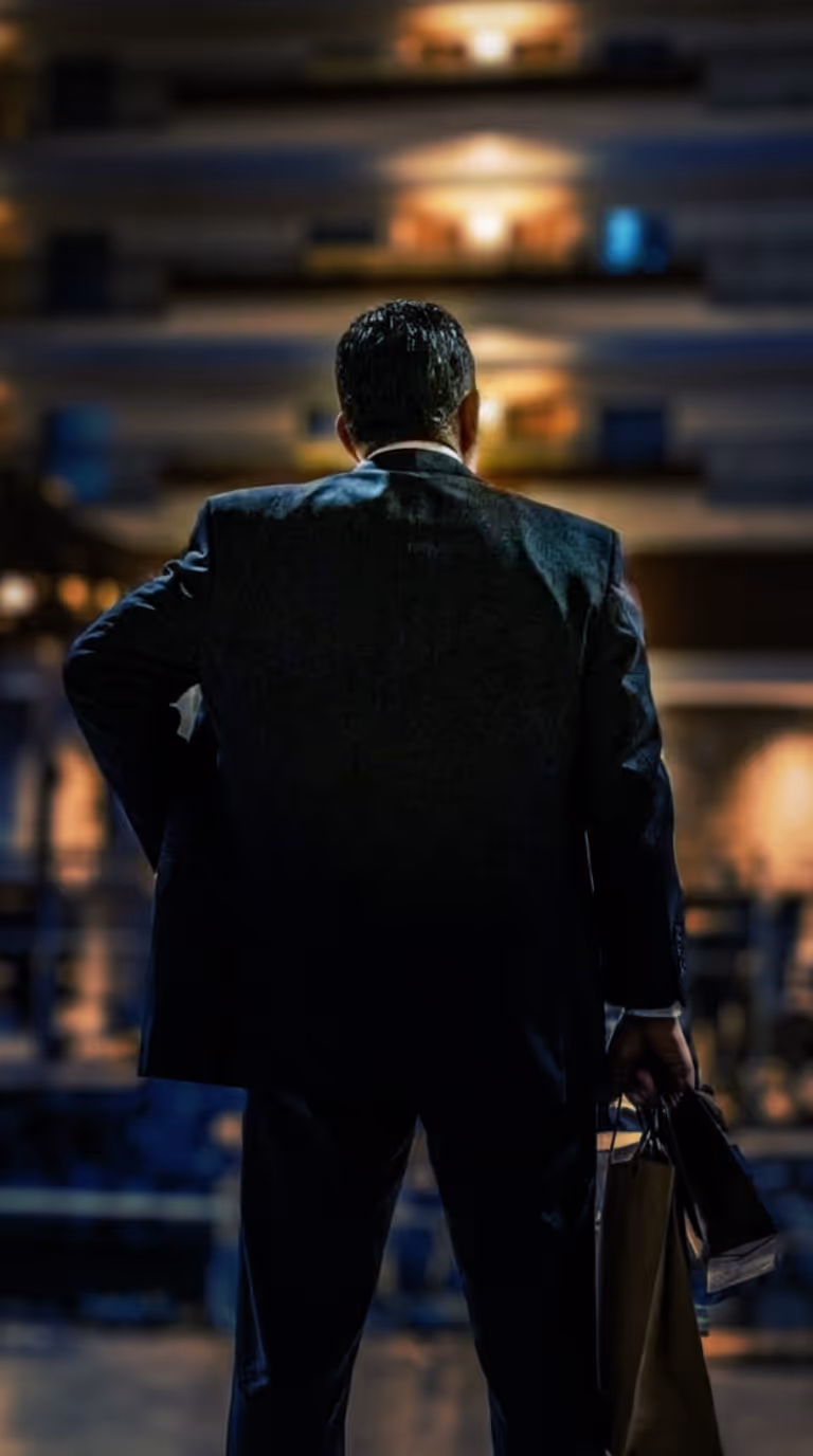 Man in a dark suit standing with his back to the camera, holding shopping bags, in a dimly lit urban environment at night.