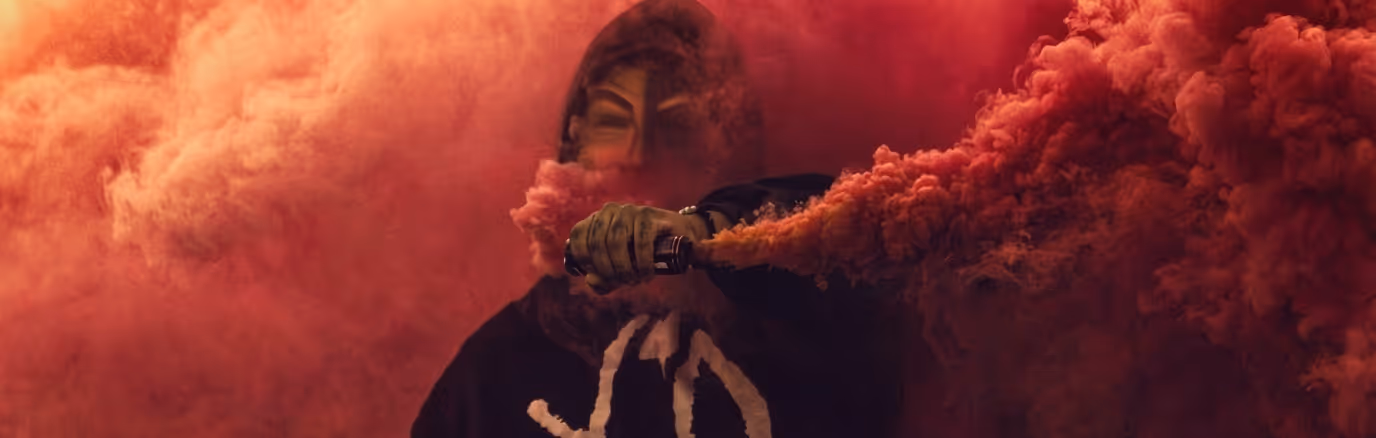 Person wearing a Guy Fawkes mask surrounded by thick red smoke while holding a red smoke flare.