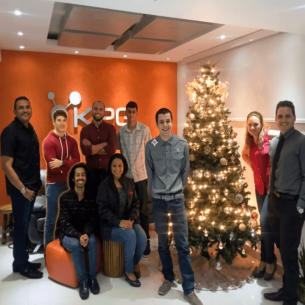 Group of eight people posing beside a lit Christmas tree in a modern office with an orange wall logo.