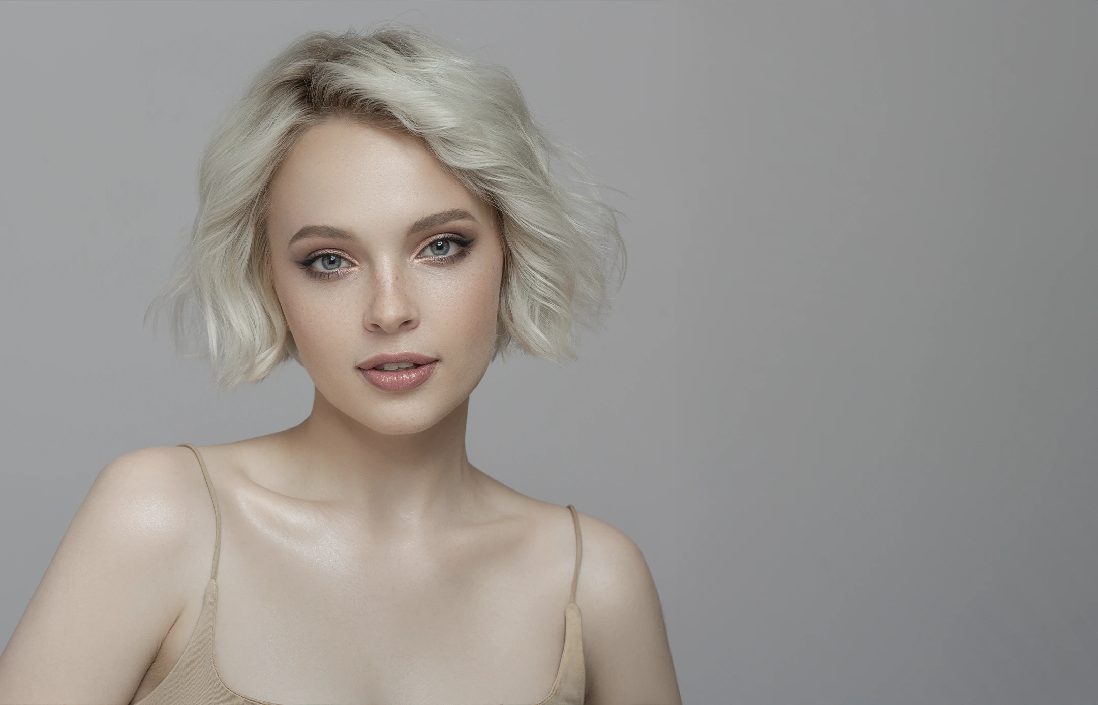 Portrait of a young woman with short platinum blonde hair and blue eyes wearing a beige spaghetti strap top against a gray background.