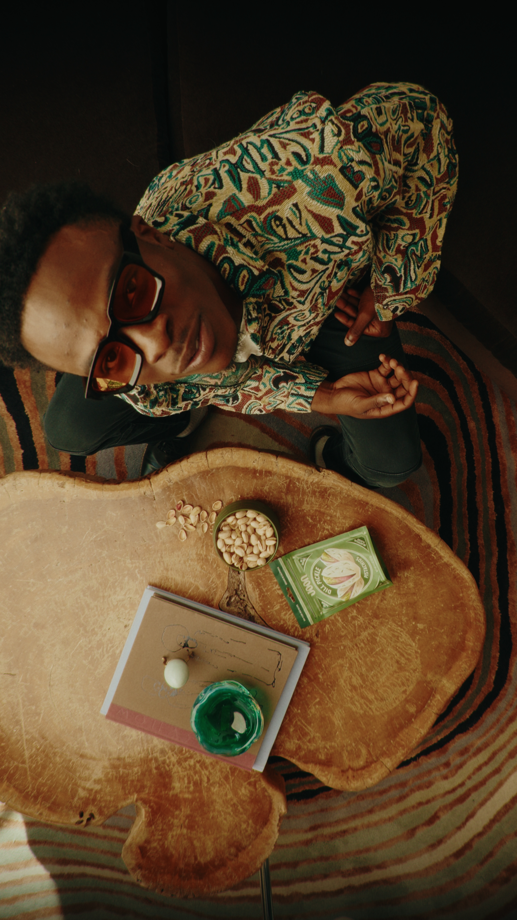Man wearing patterned jacket and tinted glasses sitting on a striped carpet beside a wooden table holding pistachios, a green cup, and notebooks.
