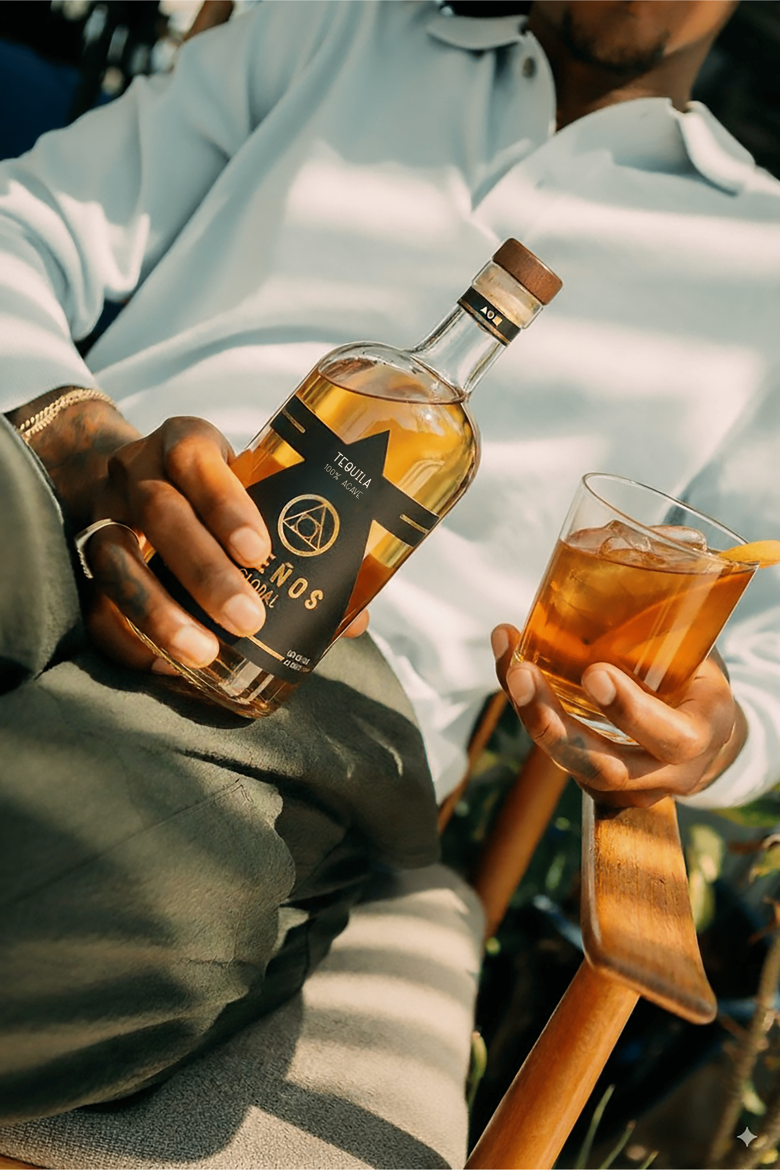 Person holding a bottle of Añejo Tequila and a glass with iced drink and a lemon slice, sitting on a wooden chair.