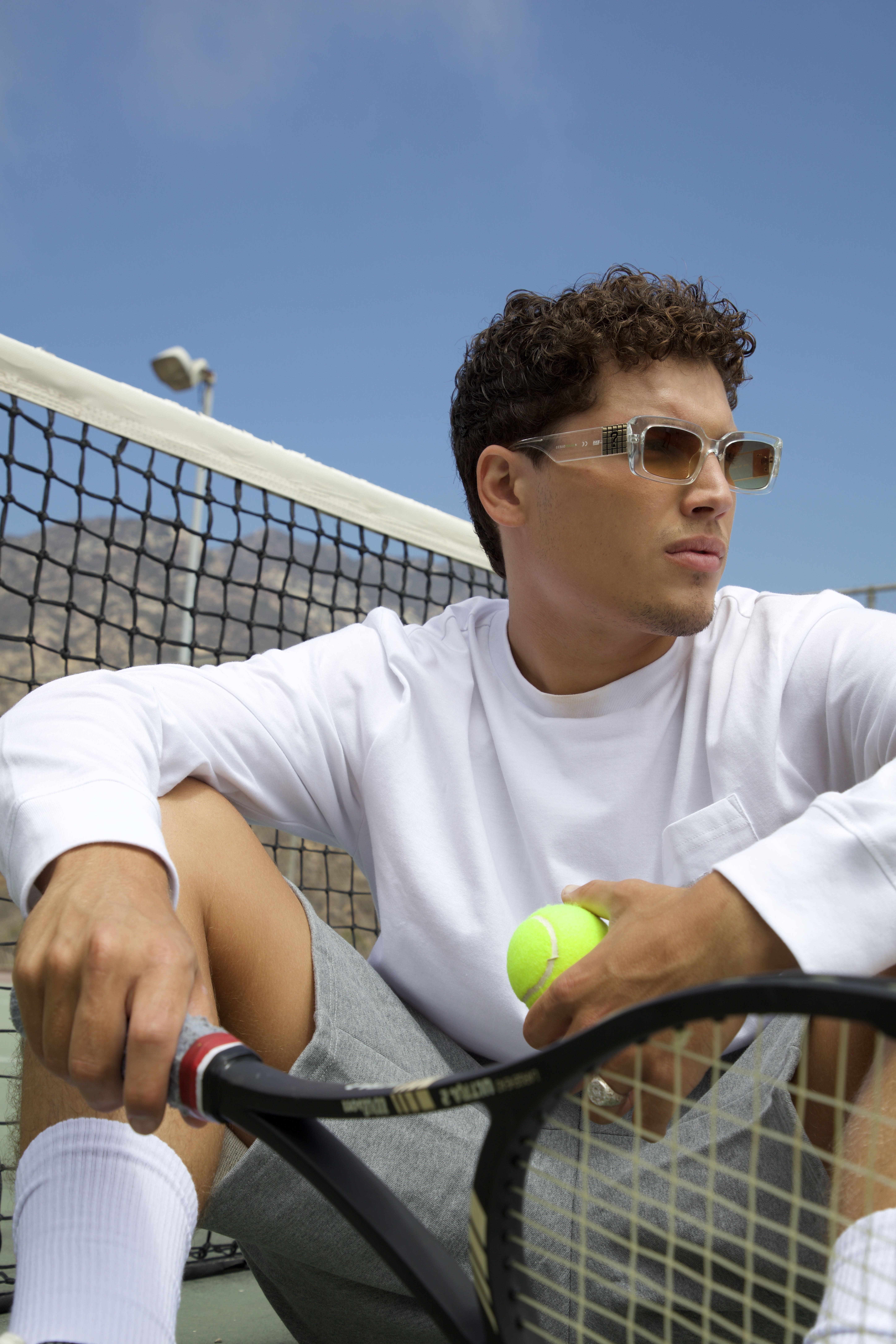 Young man wearing sunglasses holding a tennis ball and racket sitting near a tennis net under a clear blue sky.