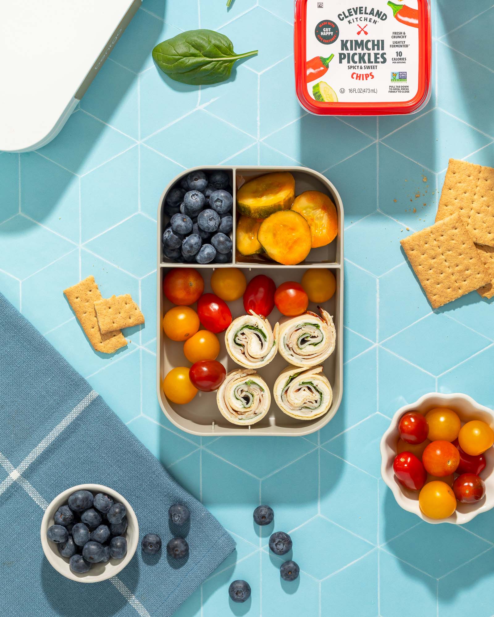 And overhead shot of a packed bento box with tomatoes, blueberries, turkey pinwheels and Cleveland Kitchen Kimchi Pickles