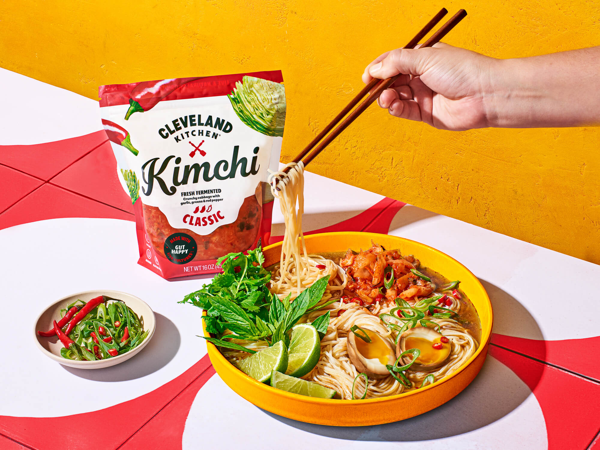 Hand using chopsticks to scoop noodles out of bowl topped with Cleveland Kitchen Classic Kimchi.