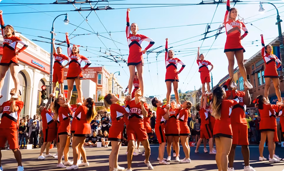Cheerleading team in red uniforms performing stunts on a city street during a sunny day with an audience watching.