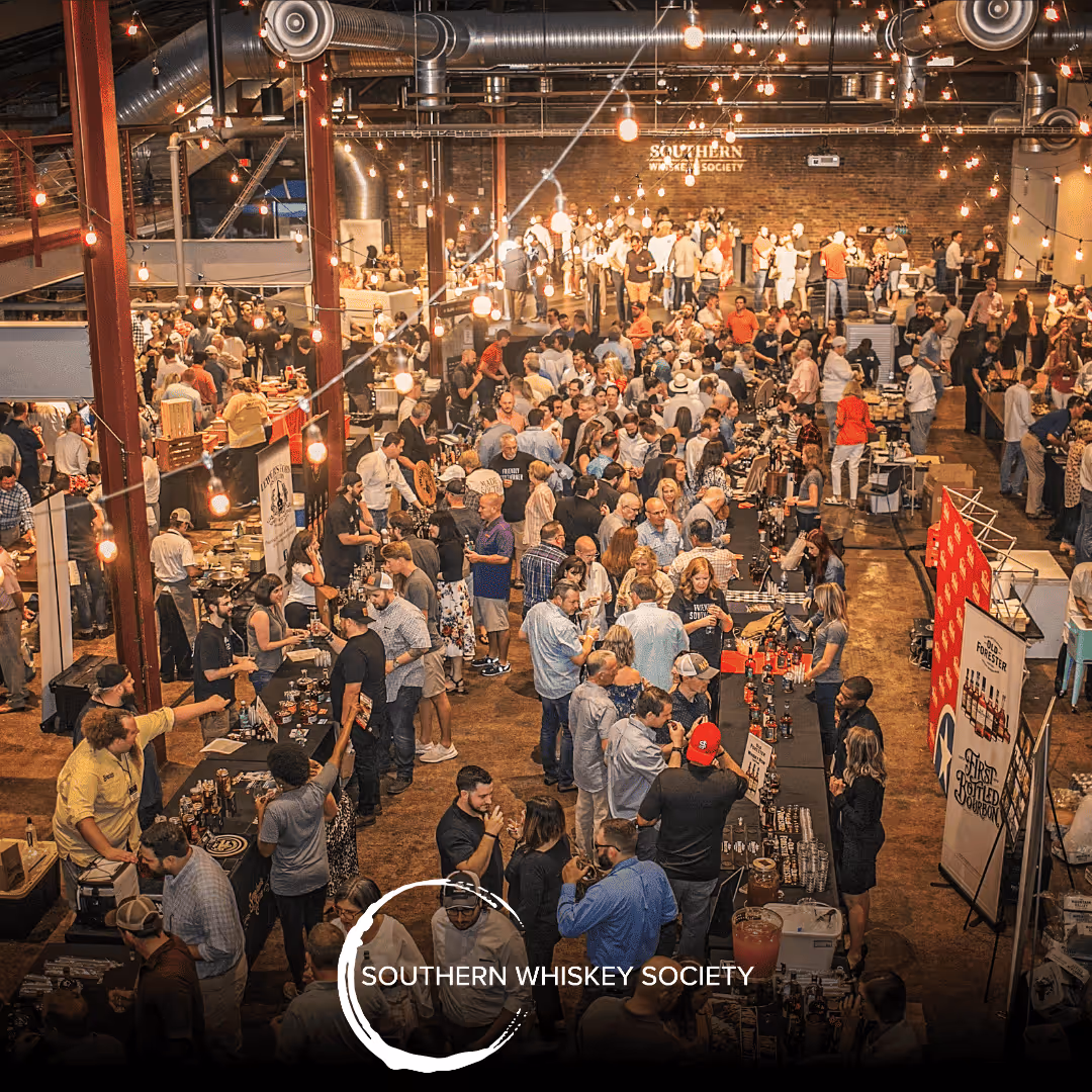 Large crowd at Southern Whiskey Society event in a spacious industrial venue with tasting tables and string lights.