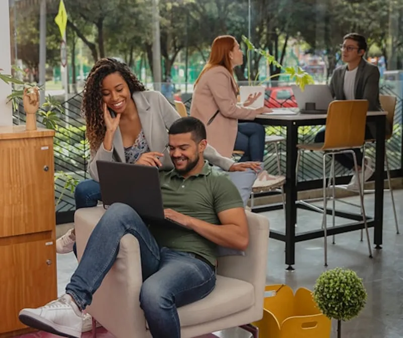 Two men and two women in a modern, bright office space; one man sits on a lounge chair using a laptop while a woman behind him smiles and watches; in the background, a man and woman sit at a high table engaged in conversation with laptops open.