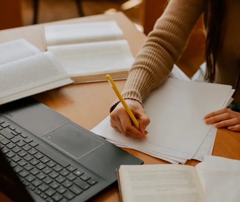 Person writing on blank papers with a yellow pencil at a wooden table surrounded by open books and a black laptop keyboard.