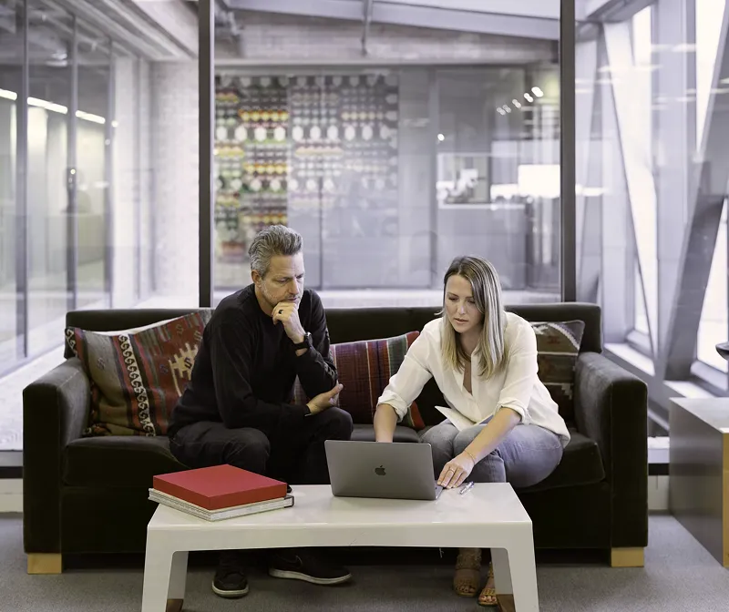 Two people sitting on a dark couch with patterned pillows, collaborating on a laptop placed on a white coffee table in front of them inside a modern office with glass walls.