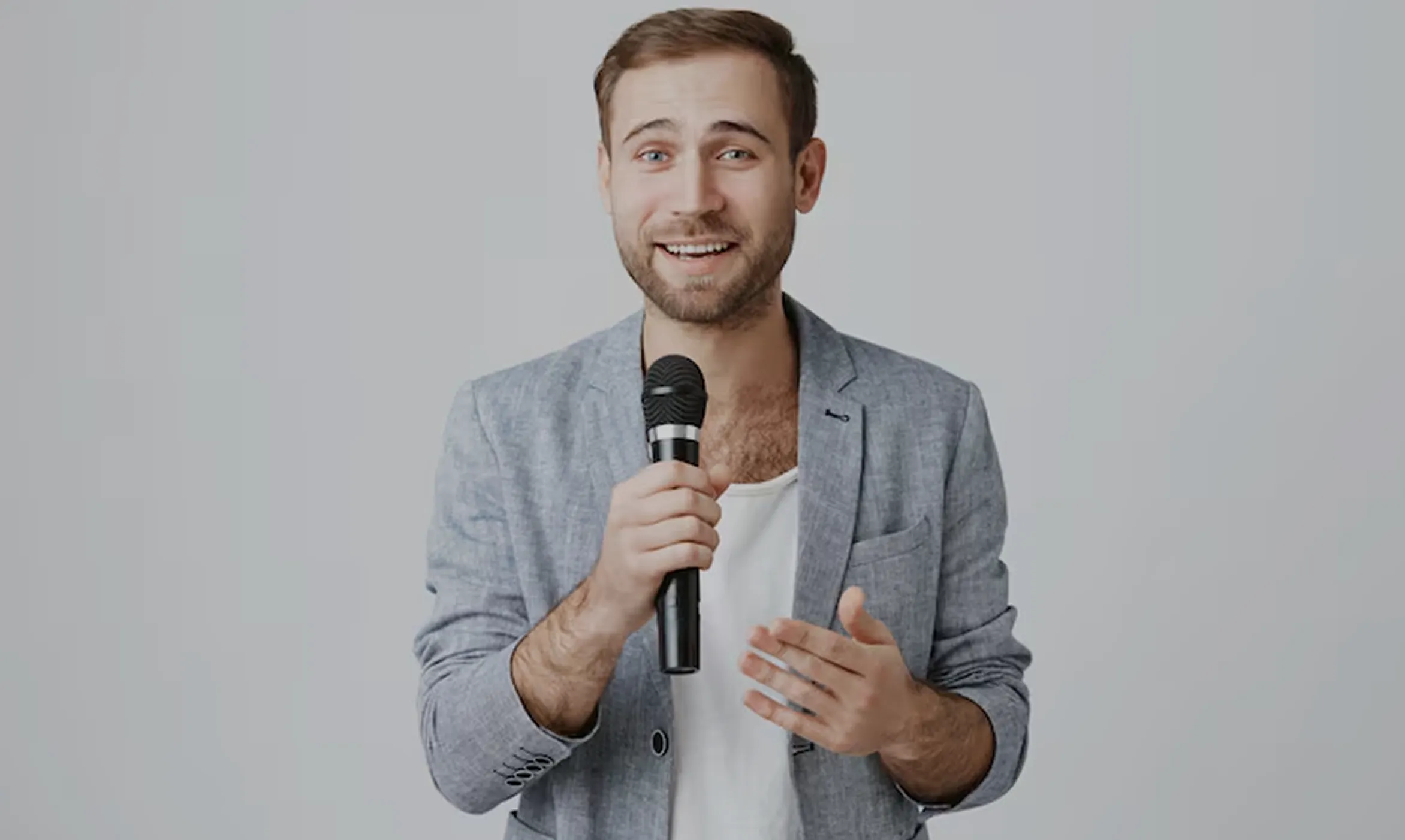A smiling man with light brown hair and beard, wearing a gray blazer and white shirt, holding a microphone and gesturing with his other hand against a plain light gray background.