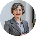Professional woman with short gray hair wearing a gray blazer and light blouse, smiling slightly and facing forward against a neutral background.