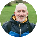 Smiling bald man with a gray beard wearing a blue and black jacket, outdoors on green grass.