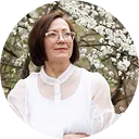 A middle-aged woman with short brown hair and glasses wearing a white blouse, standing outside in front of tree branches with white blossoms.
