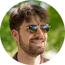 Smiling young man with a beard and curly hair wearing round reflective sunglasses and a white shirt, outdoors with green foliage in the background.