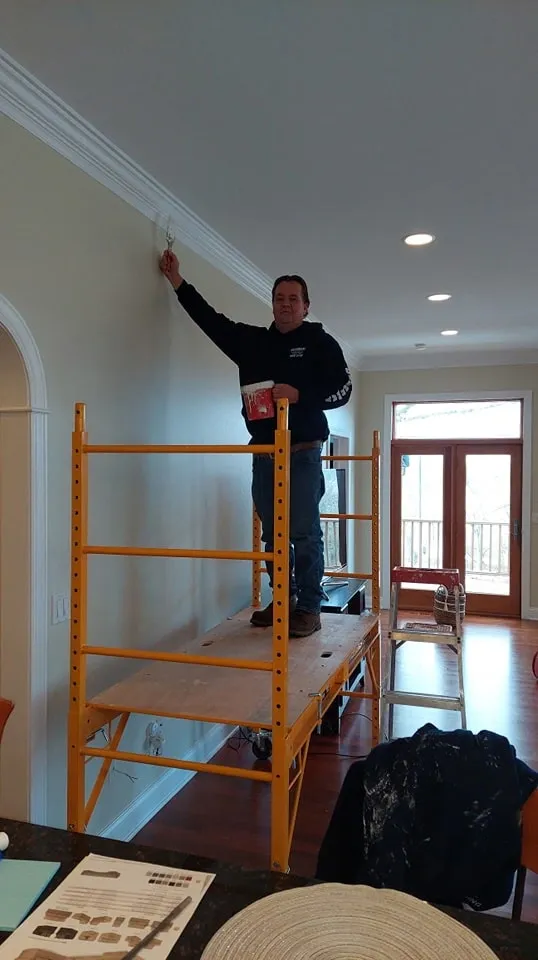 Man standing on a yellow scaffold painting the upper corner of a beige wall inside a well-lit room.