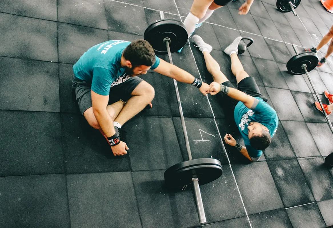 Two men in teal shirts giving a fist bump while sitting and lying on a gym floor near barbells.