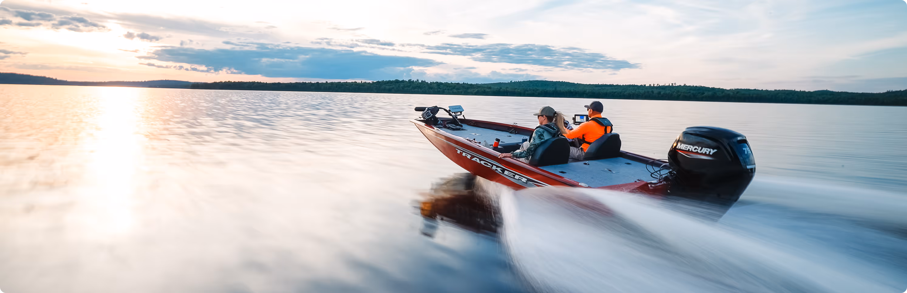 Two people riding in a red Tracker motorboat speeding across a calm lake at sunset.