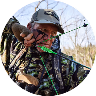 Man in camouflage clothing aiming a compound bow with a green arrow in an outdoor setting.
