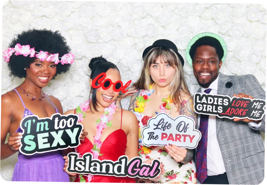 Four friends smiling at a party photo booth holding signs that say 'I'm too sexy', 'Cool', 'Island Gal', 'Life Of The Party', and 'Ladies Love Me Girls Adore Me', dressed in colorful outfits with flower leis and hats.