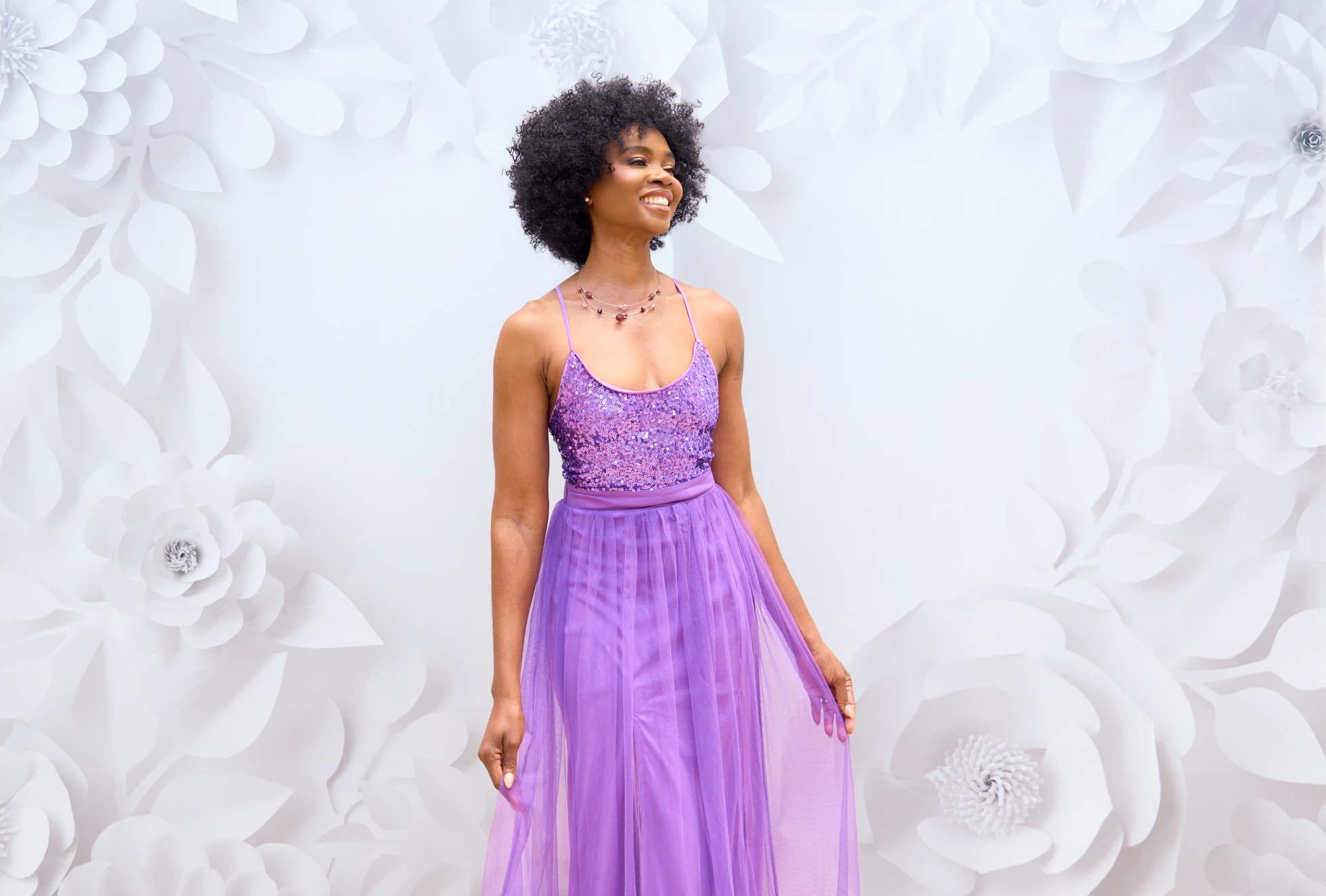 Smiling woman with curly hair wearing a purple sequin top and flowy skirt standing against a white floral backdrop.