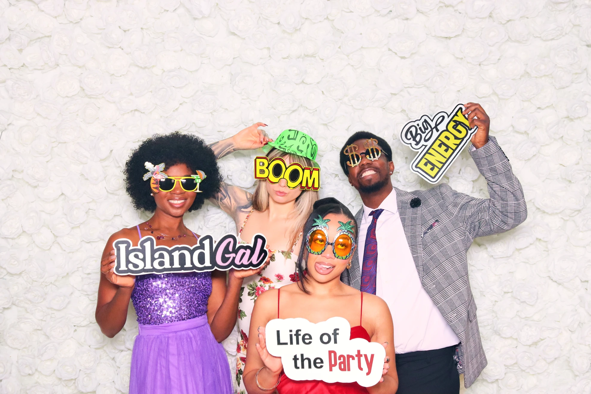 Four people dressed festively wearing novelty glasses and holding signs reading Island Gal, BOOM, Life of the Party, and Big Energy in front of a white flower wall.