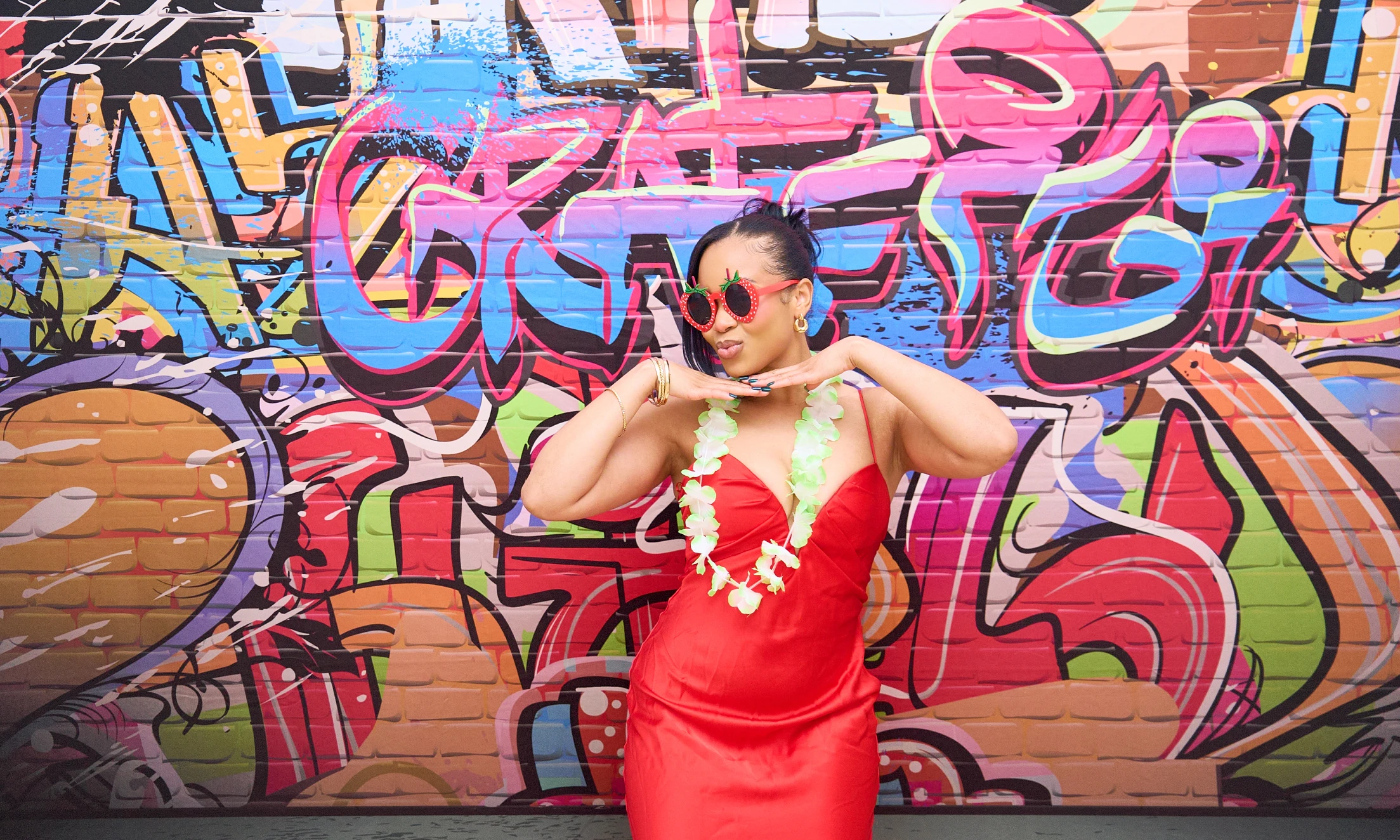 Woman in red dress with strawberry sunglasses and green lei posing playfully in front of colorful graffiti wall.
