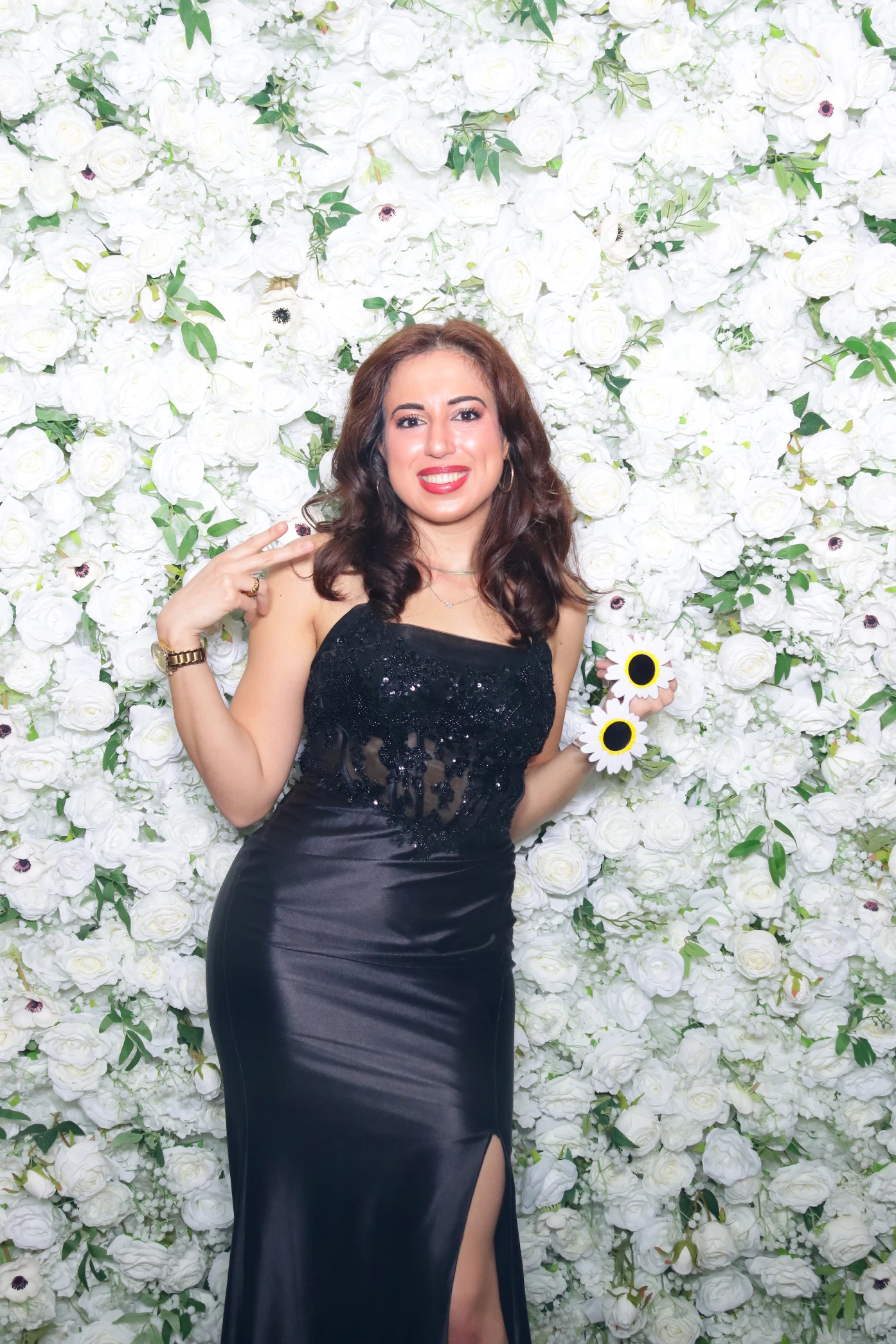 Smiling woman in a black dress posing in front of a white flower wall holding sunflower-shaped glasses.